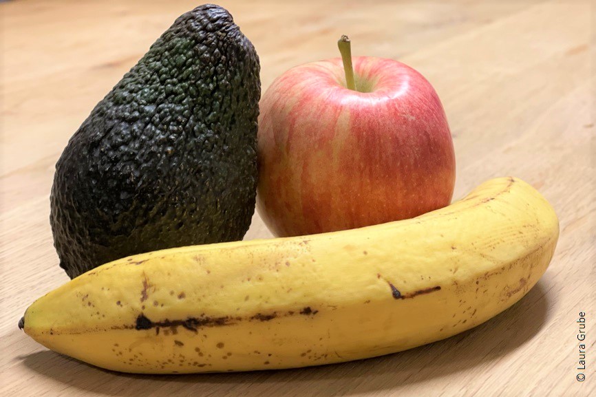 Eine Avocado, ein Apfel und eine Banane liegen auf einem Holztisch. 